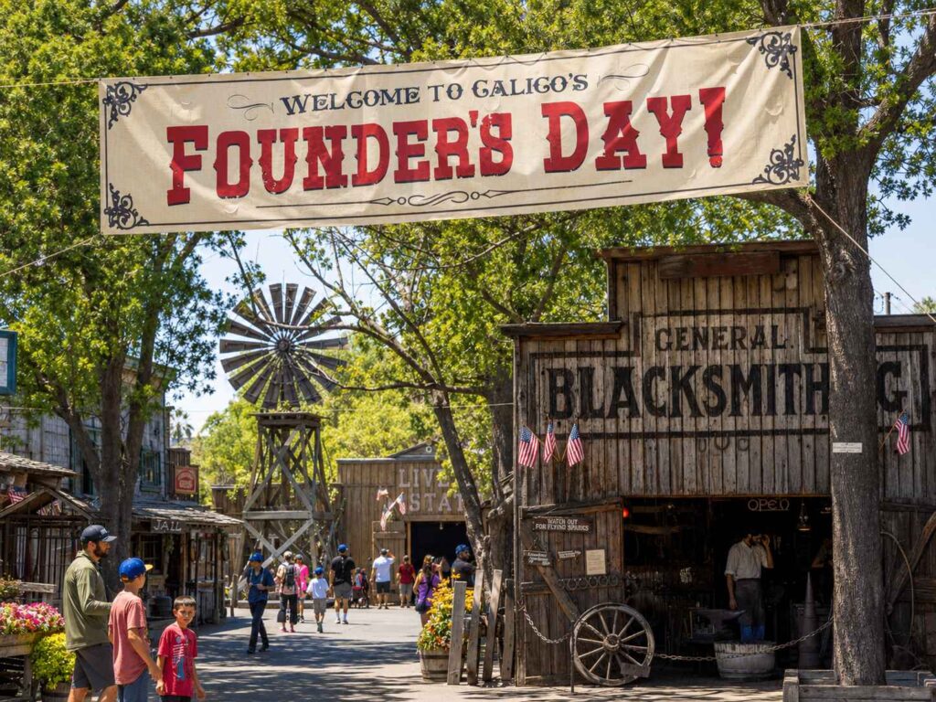 Knott's Berry Farm Ghost Town decorated for Calico's Founder's Day, with a vintage Welcome banner overhead, the General Blacksmith shop, a windmill, and families walking through the Old West street on a sunny day