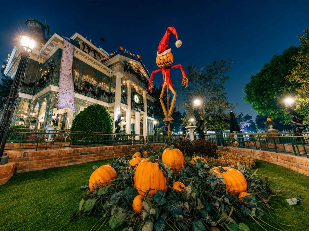 Haunted Mansion Holiday at Disneyland at night, decorated for the Nightmare Before Christmas overlay, with a jack-o-lantern scarecrow in a Santa hat standing over a pumpkin patch on the front lawn