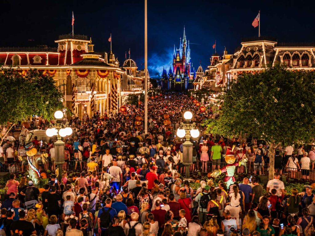 Large nighttime crowd packed along Main Street U.S.A. at Magic Kingdom, facing the illuminated blue Cinderella Castle, with Halloween pumpkins and scarecrow decorations lining the street
