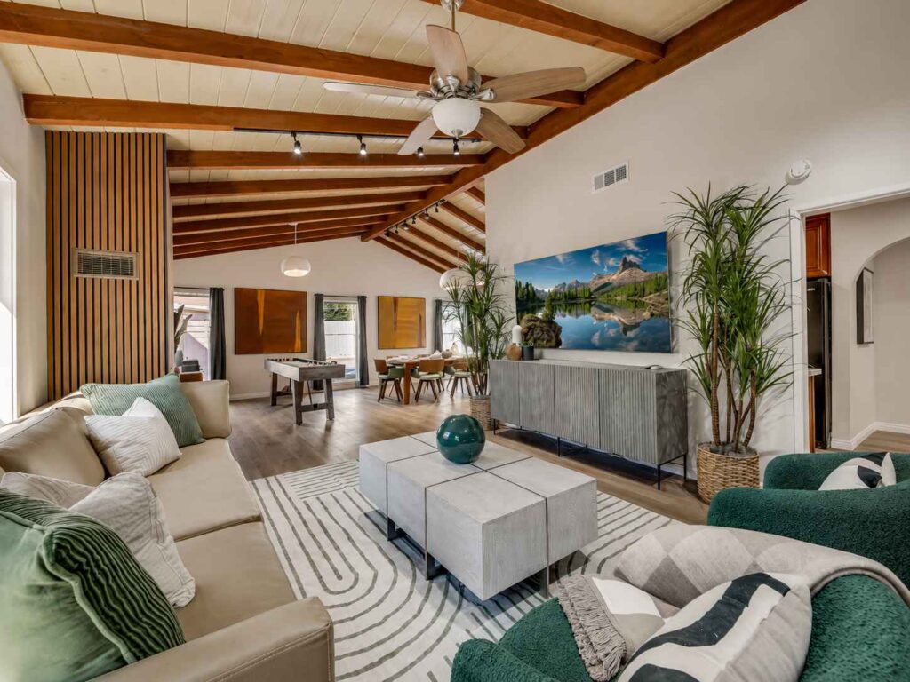 Spacious open-concept living room with a vaulted wood-beam ceiling, beige sectional and green accent chairs, large wall-mounted TV, foosball table, and dining area with natural light and indoor plants