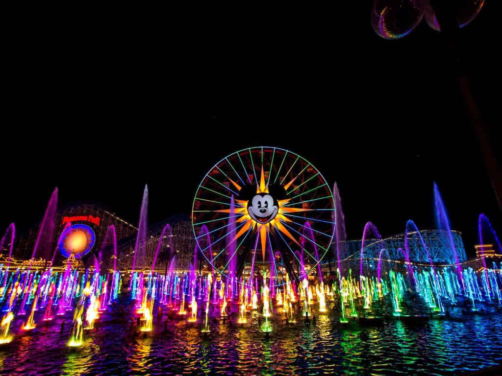 Colorful World of Color fountain show at night in Disney California Adventure, with Mickey's Fun Wheel and Paradise Pier lit up behind illuminated rainbow water jets